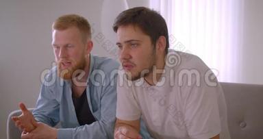 两位英俊的白种人成年男子在电视上兴奋地坐在沙发上观看体育节目的特写照片